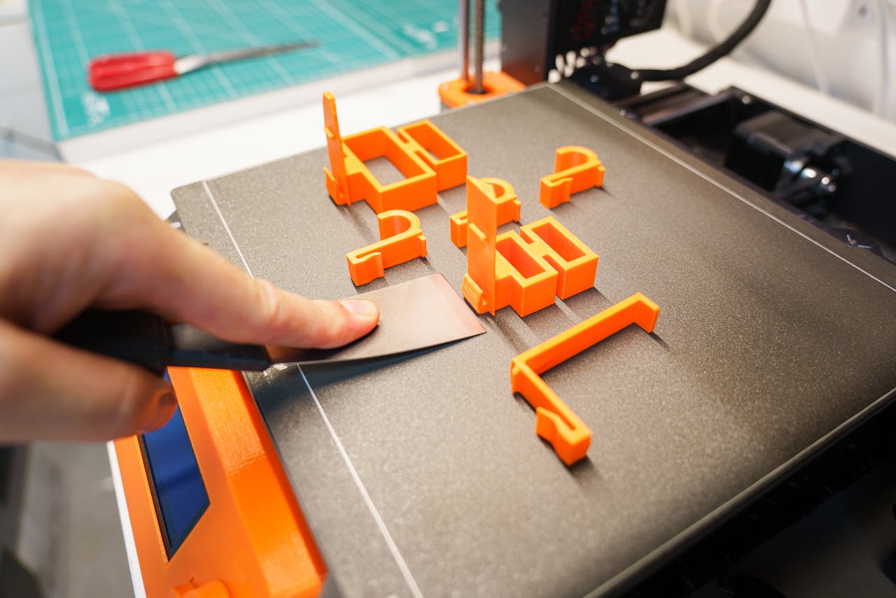 A hand carefully removes an orange 3D printed object from a printer bed using a spatula indoors.